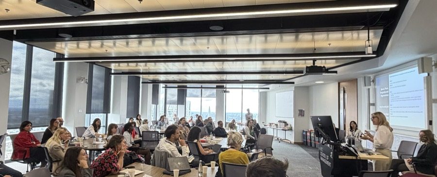 Researchers seated around tables in a top-floor room at UCL School of Management, listening to a speaker at the lectern, with the London skyline through the windows.